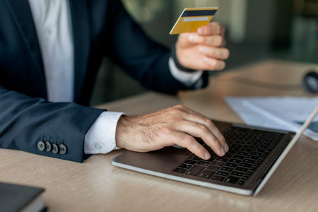 Male entrepreneur using credit card and laptop while sitting at workplace in office, focus on hand on keyboardの写真素材