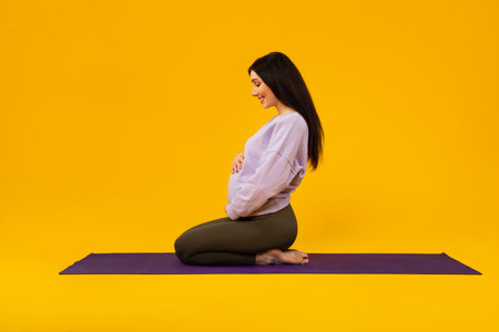 Young pregnant lady in sportswear touching belly and smiling, sitting on yoga mat over yellow...
