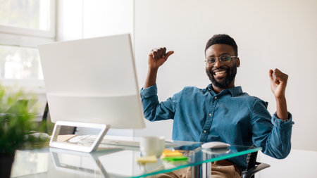 Excited black businessman gesturing yes in front of computer, celebrating success, sitting at workplace in office, panoramaの写真素材