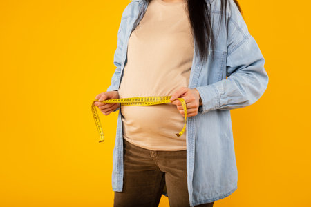 Young pregnant lady measuring her belly with tape to keep track of her fetus development, cropped,...
