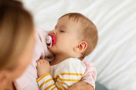 Small baby girl on mothers hands sucking pacifier and sleeping peacefully, above view, closeup
