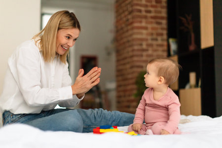 Happy mother smiling and playing clap hand with her adorable little daughter, sitting on bed at home
