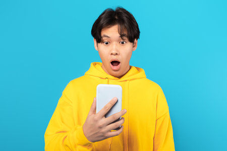 Shocked Asian Teen Boy Holding Smartphone Posing On Blue Backgroundの写真素材