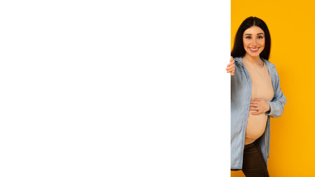 Excited pregnant lady peeking out of blank white board for text or design, yellow background, mockupの写真素材