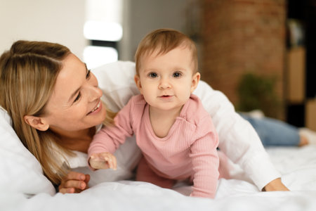Motherhood. Happy mom playing with cute toddler daughter, crawling on bed looking at camera