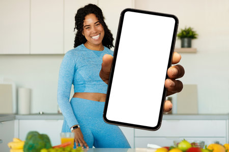 Dieting App. Black Woman In Sportswear Standing In Kitchen, Showing Blank Smartphoneの写真素材