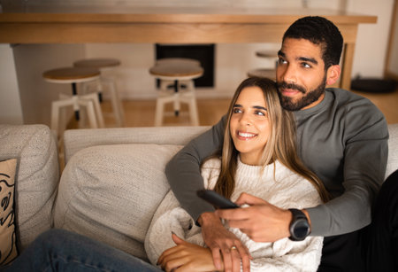 Cheerful millennial middle eastern male with beard and remote control hugs european woman, watch TVの写真素材