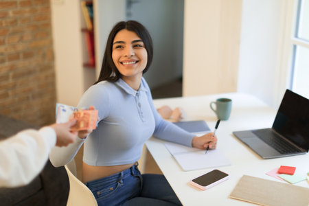 Mother giving cash to her teen spanish daughter for daily expenses, lady sitting at table and studying via laptopの写真素材