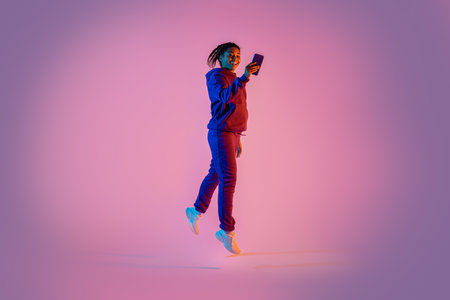 Full body length of young sporty black woman in sportswear jumping and using mobile cellphone on pink neon backgroundの写真素材