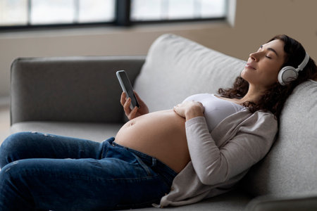 Calm Pregnant Lady In Wireless Headphones Listening Music On Smartphone At Homeの写真素材