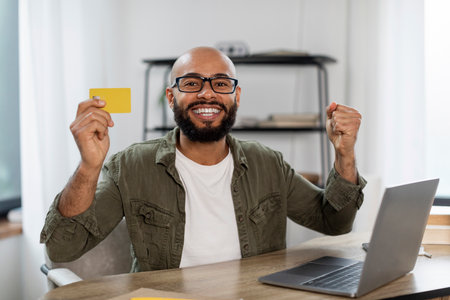 Overjoyed latin businessman holding credit card and clenching fists, smiling at camera in front of laptop at home officeの写真素材