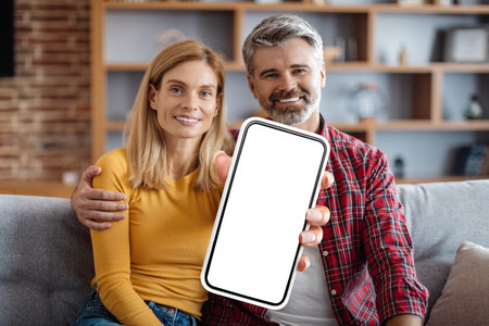 Middle Aged Couple Sitting On Couch And Showing Blank Smartphone At Camera
