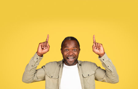Glad excited mature black guy in casual point fingers at blank space for text, isolated on yellow backgroundの写真素材