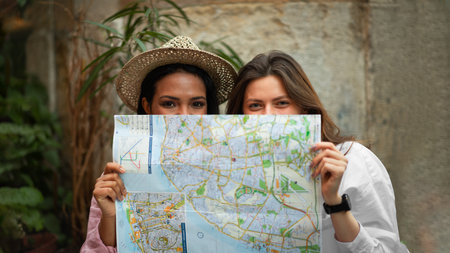 Smiling young african american and european women in hat looking for route peeking out from behind mapの写真素材