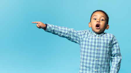 Look here. Surprised black boy pointing aside at free copy space, looking at camera with open mouth over blue backgroundの写真素材