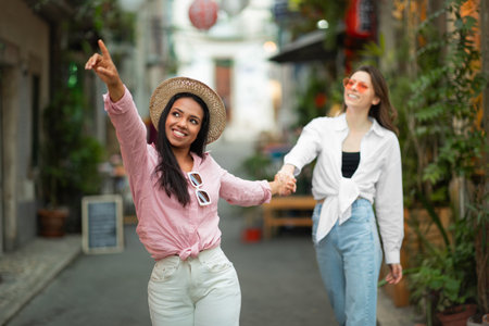 Smiling inspired millennial african american lady leads by hand european woman tourist in casual and sunglassesの写真素材