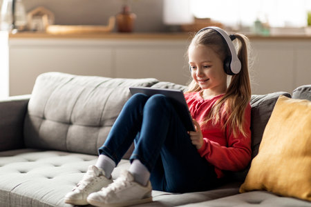 Cute Little Girl Wearing Wireless Headphones Using Digital Tablet At Homeの写真素材