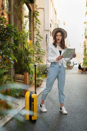 Shocked millennial european woman tourist in hat with suitcase with map looking for travel, route and hotelの写真素材