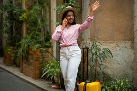 Cheerful millennial african american woman tourist in hat with suitcase calling by phone, waving hand, enjoy travelの写真素材