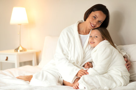 Happy european millennial woman and little girl in bathrobes hugging, sit on soft bed, enjoy rest together, free timeの写真素材