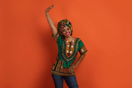 Positive funny black woman in african costume dancing on orangeの写真素材