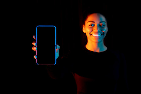 Happy black woman inviting for shopping, showing cellphone with blank screen over dark background in neon light, mockupの写真素材
