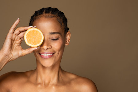 Face acids concept. Black middle aged woman using fresh juicy fruit for skin care, brown background, free spaceの写真素材