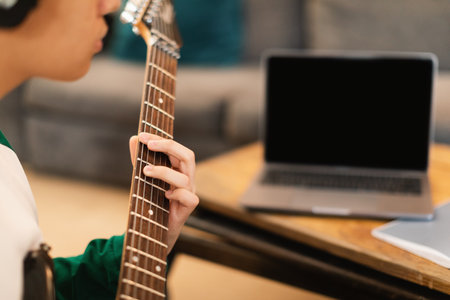 Guy Wearing Earphones Playing Electric Guitar Sitting At Laptop Indoorsの写真素材
