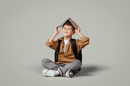 Glad little european boy with backpack puts book on head, sits on floor, enjoy learningの写真素材