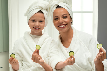 Happy european millennial lady and little girl in bathrobes and towel hold pieces of cucumber, enjoy weekendの写真素材
