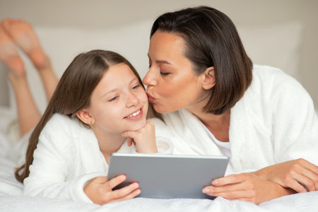 Smiling young lady kiss on cheek little caucasian girl in bathrobe, watch video on tablet, relaxing on bedの写真素材