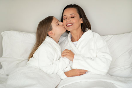 Smiling little caucasian girl kiss on cheek of young woman in bathrobe, relaxing on white soft bedの写真素材