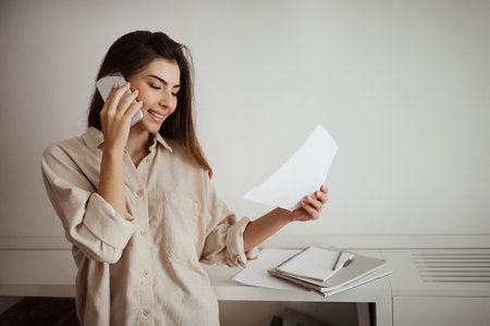 Smiling millennial mixed race woman calling by smartphone, talking with client, looking at documentsの写真素材