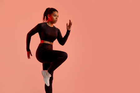 Athletic african american lady warming up before running, training on peach neon studio background,...