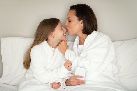 Glad young woman kissing nose little caucasian girl in bathrobe, relaxing on white soft bed, enjoy free timeの写真素材