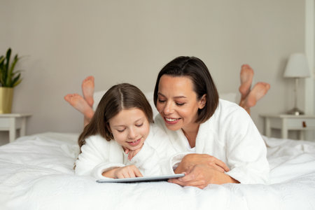 Glad young caucasian female, teen girl in bathrobe, watch video on tablet, relaxing on white soft bedの写真素材
