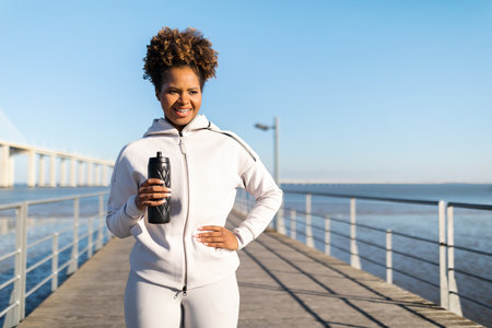 Sporty Black Female With Water Bottle In Hands Standing On Pier Outdoorsの写真素材