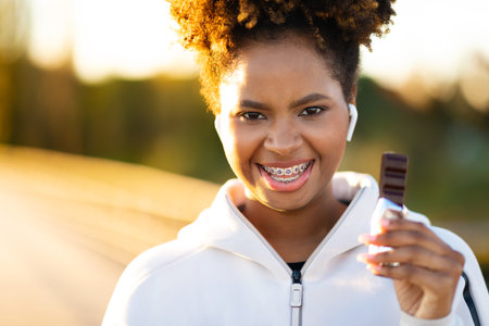 Smiling Young Black Female In Sportswear Holding Protein Bar While Standing Outdoorsの写真素材