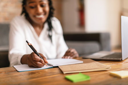 Happy black woman writing in notepad and using laptop, noting information from internet, selective...