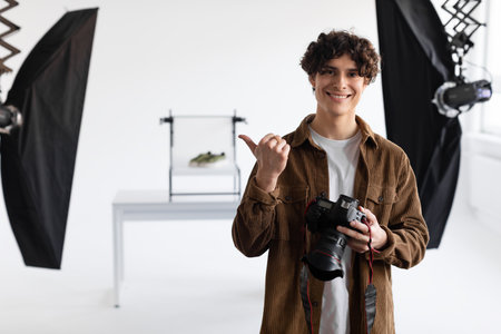 Happy young male photographer working at modern photostudio, pointing to the side, enjoying making content photoshootの写真素材
