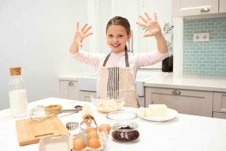 Glad caucasian small girl in apron making cookie dough, has fun, showing hands in flour in kitchen interiorの写真素材