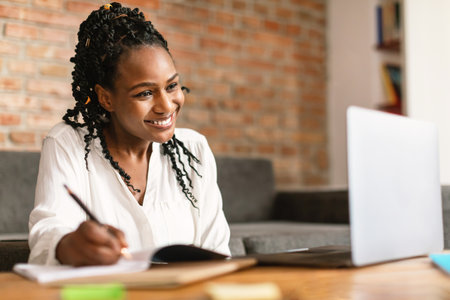 Excited black woman taking notes and using laptop, making video call while working or learning remotely from homeの写真素材