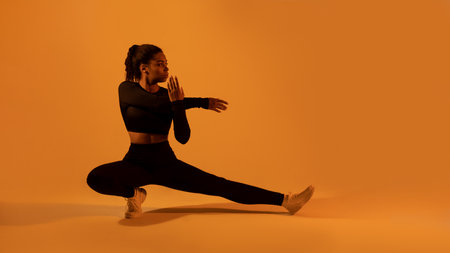 Fit black lady in sportswear stretching legs and arms, warming up, orange neon background, panorama, copy spaceの写真素材