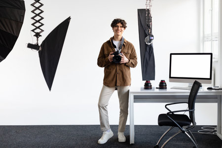 Excited male photographer with dslr camera sitting on his desk with computer monitor with white empty screen, templateの写真素材