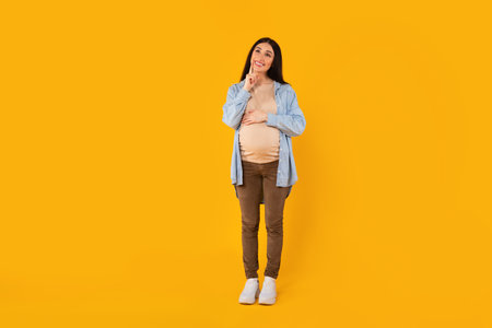 Happy pensive pregnant woman touching belly and chin, looking up and thinking, standing on yellow studio backgroundの写真素材
