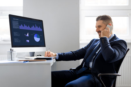 Confident middle aged businessman working on computer with graphics and charts and having business call in officeの写真素材