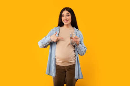 Happy pregnant woman holding glass of water and pointing at it, smiling at camera on yellow studio backgroundの写真素材