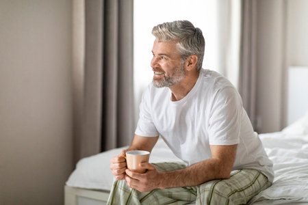 Joyful middle aged man sitting on bed, drinking coffeeの写真素材