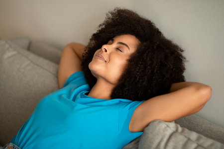 Cheerful millennial african american curly lady with closed eyes enjoys sleeping on sofa in living roomの写真素材
