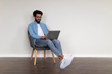 Young Indian Freelancer Guy Using Laptop While Sitting In Armchair At Homeの写真素材